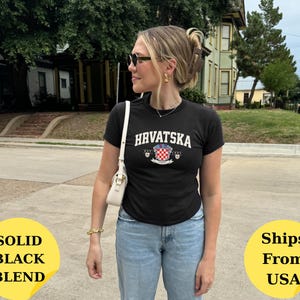 May include: A black short-sleeved t-shirt featuring the white text "HRVATSKA" in an arched font above a red and white checkered crest emblem. The shirt is paired with light blue denim jeans and a small white shoulder bag with gold hardware.
