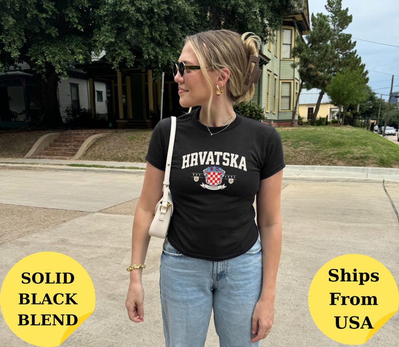 May include: A black short-sleeved t-shirt featuring the white text "HRVATSKA" in an arched font above a red and white checkered crest emblem. The shirt is paired with light blue denim jeans and a small white shoulder bag with gold hardware.