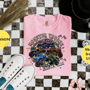 Peut inclure: T-shirt rose clair avec un motif de monster trucks multicolores et le texte "Monster Trucks Are My Jam". L'image comprend des baskets blanches et des &eacute;l&eacute;ments d&eacute;coratifs sur un fond &agrave; carreaux noir et blanc.