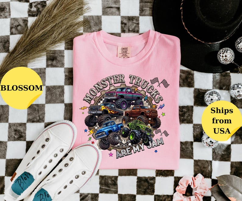 Peut inclure: T-shirt rose clair avec un motif de monster trucks multicolores et le texte "Monster Trucks Are My Jam". L'image comprend des baskets blanches et des &eacute;l&eacute;ments d&eacute;coratifs sur un fond &agrave; carreaux noir et blanc.