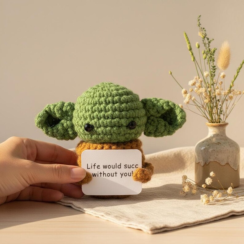 May include: A handmade green crochet figure with large ears, holding a card that reads "Life would succ without you!". The figure has black eyes and a tan body. A small vase with dried flowers sits to the right.
