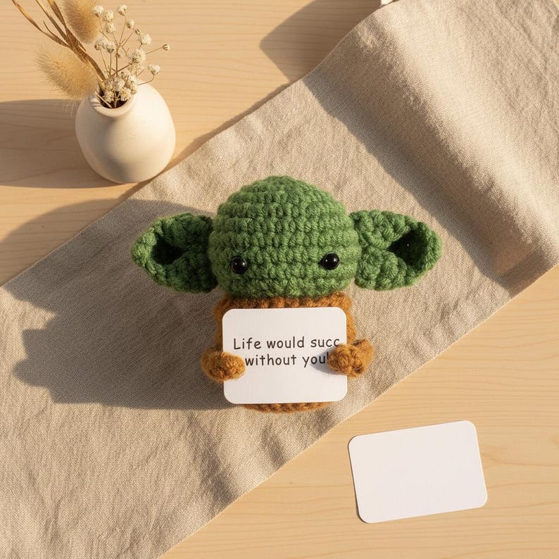 May include: A green and brown crocheted figure with large ears, holding a white card with the text "Life would succ without you!". The figure is placed on a beige cloth, with a small white vase and dried flowers in the background.