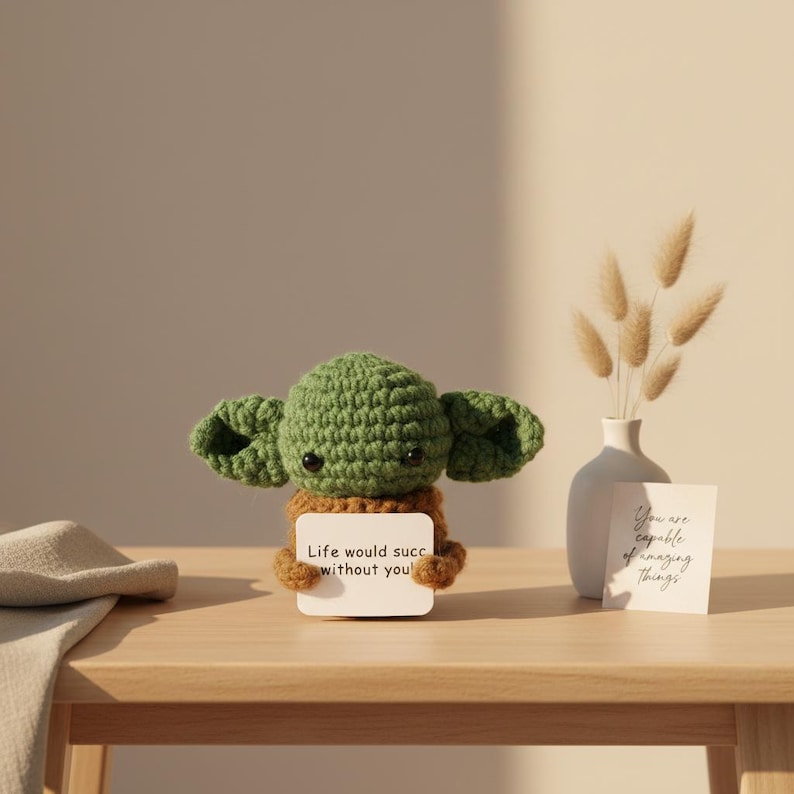 May include: A crocheted green and brown figure with large ears, holding a sign that reads "Life would succ without you." A small white vase with dried flowers and a card that says "You are capable of amazing things" are also on the wooden table.