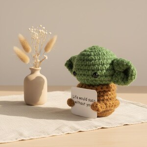 May include: A handmade green and brown crocheted figure holding a small white card with the text "Life would succ without you." A small beige vase with dried flowers is to the left.