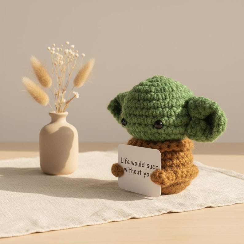 May include: A handmade green and brown crocheted figure holding a small white card with the text "Life would succ without you." A small beige vase with dried flowers is to the left.