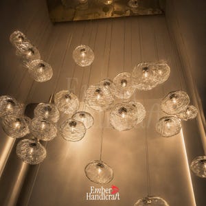 May include: A multi-pendant chandelier featuring numerous clear, textured glass globes suspended at varying heights. Each orb contains a warm light source, creating a cluster of illuminated spheres. The text Ember Handicraft is visible at the bottom.