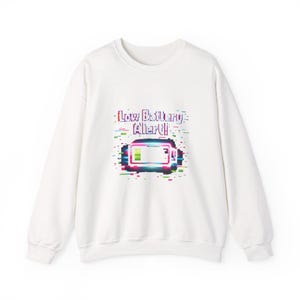May include: White sweatshirt with a graphic that reads "Low Battery Alert!" in a pixelated style. The battery icon is a rectangle with a green and pink fill, and the text and icon have a glitch effect.