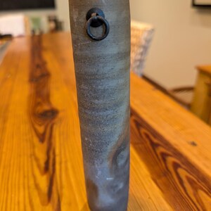 May include: A tall, cylindrical ceramic vase with a textured, mottled gray and brown finish. A small, dark metal ring is attached near the top. The vase is displayed on a wooden surface.