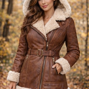 May include: A brown jacket with a cream-colored faux shearling lining, a hood, and a belted waist. The jacket features a front zipper closure, two zippered side pockets, and faux shearling trim on the hood, cuffs, and bottom hem.