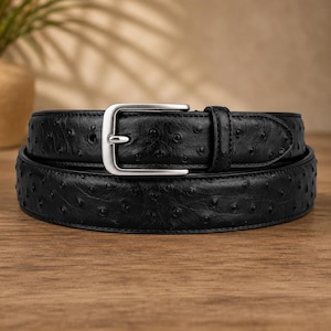 Puede incluir: Un cinturón negro con un patrón texturizado estilo avestruz que presenta protuberancias en relieve por toda la superficie. El cinturón incluye una hebilla de metal plateado pulido con una sola púa y una trabilla de cuero negro a juego.