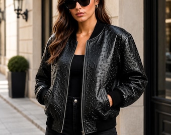 May include: A black bomber jacket featuring a textured, dotted surface pattern, a front zipper closure, and solid black ribbed cuffs and collar. The jacket is worn over a plain black top and paired with black pants, complemented by dark sunglasses.