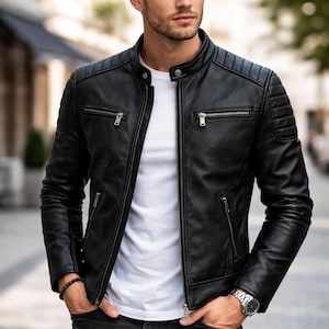 May include: A black leather jacket with a stand-up collar, silver-tone metal zippers, and quilted shoulder detailing. The jacket is worn over a plain white t-shirt and paired with dark grey denim jeans, featuring a beaded bracelet and a metal watch.