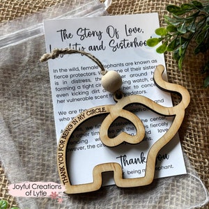 May include: Wooden elephant ornament with a heart cutout and the text "Thank you for being in my circle" engraved on it. The ornament is hanging from a string with a wooden bead. The ornament is on a white background with a piece of brown burlap fabric.