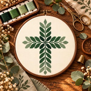 Peut inclure: Un tambour à broder circulaire en bois contenant un motif de point de croix représentant une croix formée de feuilles stylisées dans des nuances variées de vert foncé et clair sur un tissu de couleur crème, centré dans le cadre.