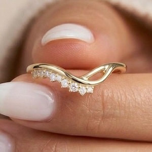 Op de afbeelding: Een goudkleurige ring met een gebogen ontwerp, bezet met kleine, heldere edelstenen. De ring wordt tussen de vingers gehouden en toont zijn delicate details en elegante stijl. De ring is een sieraad.