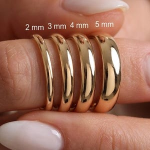 Puede incluir: Cinco anillos dorados de diferentes anchos, que van de 2 mm a 5 mm, se muestran en un dedo. Los anillos tienen un acabado brillante y pulido. La imagen incluye el texto "2 mm 3 mm 4 mm 5 mm". Estos anillos son una joya.