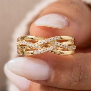 May include: A gold-colored ring with a woven design. The ring features multiple bands that intersect, creating an intricate pattern. Small, clear stones are set along the bands, adding sparkle. The ring is held by a person with manicured nails.