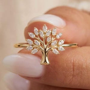 May include: A gold ring with a tree motif. The tree's leaves are made of small, clear gemstones. The ring is held up by a person's hand, highlighting the ring's intricate design and craftsmanship. The ring is a piece of jewellery.