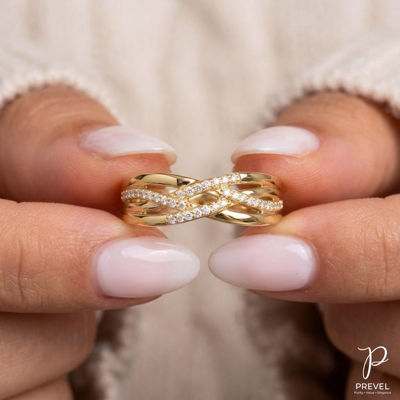 May include: A gold-colored ring with a woven design, encrusted with small, sparkling gemstones. The ring is held between two hands with manicured nails. The ring's design features interwoven bands, creating a unique and elegant look.