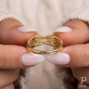 May include: A gold ring with an interwoven design, held between two hands. The ring features a series of small, sparkling accents. The ring's design is intricate and detailed, showcasing a blend of elegance and craftsmanship. The ring is a piece of jewelry.