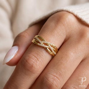 May include: A gold-colored ring with an interwoven design, adorned with small, sparkling stones. The ring features a braided pattern across the top, with multiple bands. The ring is on a finger.