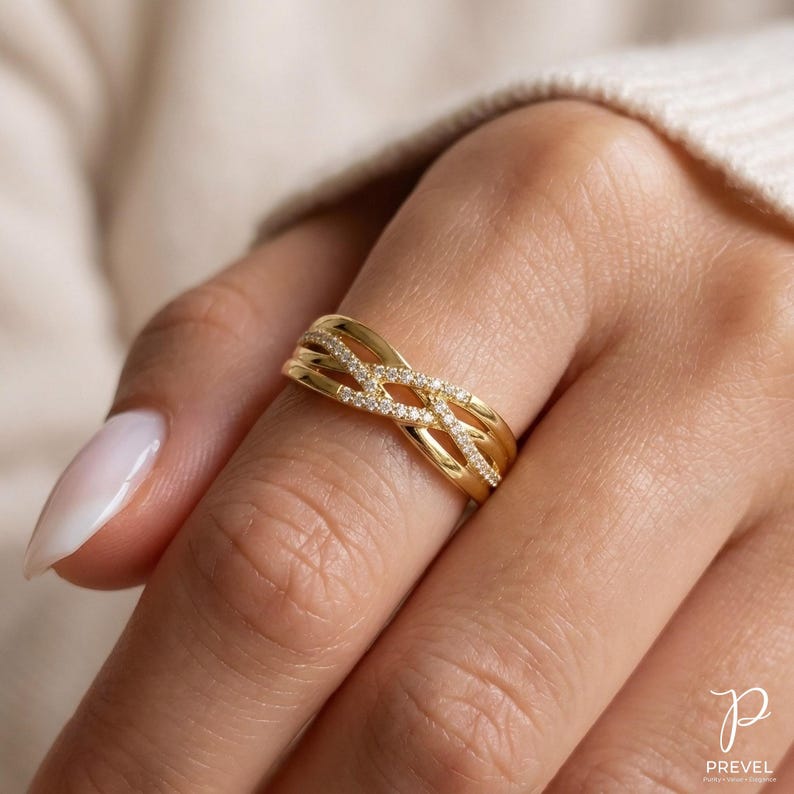 May include: A gold-colored ring with an interwoven design, adorned with small, sparkling stones. The ring features a braided pattern across the top, with multiple bands. The ring is on a finger.