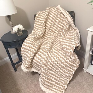 May include: A beige and white chunky knit blanket draped over a chair. The blanket features a chevron pattern and a thick, white border. The cozy throw is made of a soft, plush material, perfect for snuggling up on the couch.