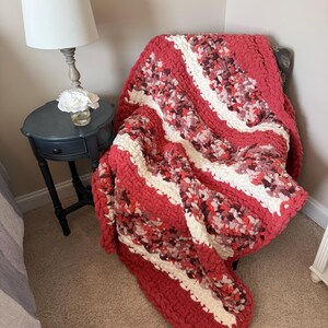 May include: A chunky knit blanket in shades of red, white, and burgundy is draped over a chair. The blanket features a striped design with a thick, textured appearance. The cosy throw is a decorative accent for a bedroom or living room.