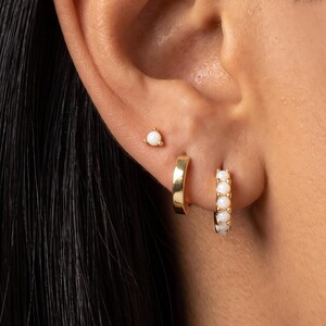 May include: A gold stud earring with a single white opal, a plain gold hoop earring, and a gold hoop earring featuring a row of small white opals. These three earrings are worn in the earlobe, showcasing a polished metallic finish and stones.