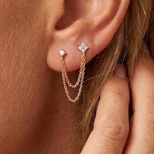 May include: A gold double-stud earring featuring two distinct points connected by a delicate gold chain. One stud is a small round stone, while the other is a four-pointed star shape set with a clear stone. The chain drapes between the two studs.