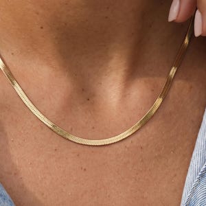 Könnte beinhalten: Eine goldfarbene Fischgräten-Halskette, die am Hals anliegt. Die Kette zeichnet sich durch ein flaches, flexibles Glieddesign aus, das eine glatte, durchgehende metallische Oberfläche erzeugt. Sie wird zu einer hellblauen Jeansjacke getragen.