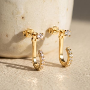May include: A pair of gold-toned earrings featuring a J-shaped design. Each earring has a single clear round stone at the top post and a row of small clear stones embedded along the curved lower section of the metal frame.