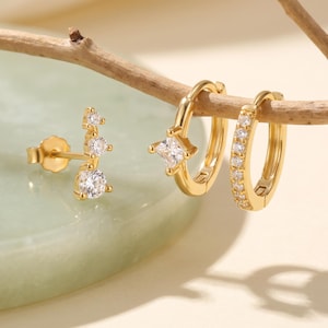 May include: A set of three gold-toned earrings featuring clear, sparkling stones. One earring is a stud with three graduated round stones, while the other two are hoop earrings, one with a single square stone and one with a row of small round stones.