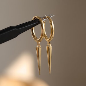 Könnte beinhalten: Ein Paar goldfarbene Creolen mit polierter Oberfläche. Jeder Ring ist mit einem abnehmbaren, schlanken, kegelförmigen Anhänger versehen, der vertikal an der Unterseite des kreisförmigen Rings hängt und ein glattes und einheitliches Erscheinungsbild erzeugt.