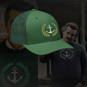 Könnte beinhalten: Grüne Trucker-Cap mit einem weißen Anker-Emblem in einem Lorbeerkranz. Die Kappe hat einen Mesh-Rücken und einen gebogenen Schirm. Das Anker-Emblem ist in Weiß, Rot und Grün gestickt.
