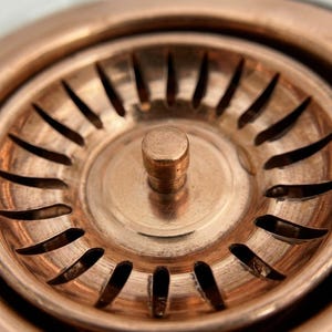 May include: A circular copper-toned sink strainer featuring a central cylindrical knob and a series of evenly spaced, elongated radial slots arranged in a ring pattern around the centre, designed for filtering debris in a kitchen sink drain.