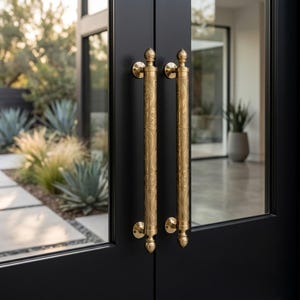 Pode incluir: Dois puxadores de porta verticais em latão com padrões gravados intrincados montados numa porta dupla preta mate. Cada puxador apresenta um corpo cilíndrico com remates decorativos no topo e na base, fixados por placas circulares à porta.