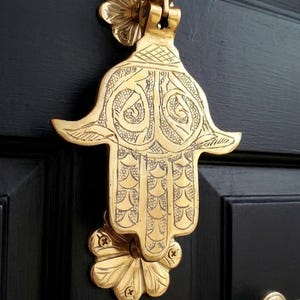 Pode incluir: Um aldravão de porta em latão polido em forma de mão de hamsa, com padrões gravados intrincados e uma placa de montagem em forma de flor no topo e na base. A superfície metálica tem um tom dourado refletor sobre uma porta preta.