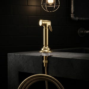 May include: A brushed gold side spray kitchen tap attachment featuring a cylindrical head, a curved handle lever, and a flexible metal hose. The unit is mounted through a dark surface with a white plastic nut visible at the base of the fixture.