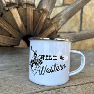 Op de afbeelding: Witte emaille campingmok met een zwart-witte illustratie van een cowboy die een paard berijdt. De mok heeft een zwarte rand en een zwarte handgreep en staat "Wild & Western" in een zwart handschrift.