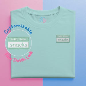 Può includere: T-shirt azzurra con una finta toppa che dice "hello, I have snacks". La toppa è verde chiaro con testo bianco e una striscia bianca. La maglietta ha uno scollo tondo e un piccolo logo vicino allo scollo.