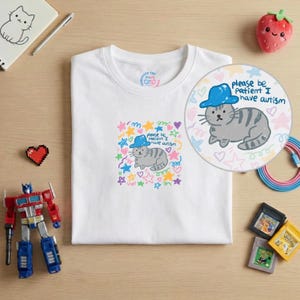 May include: White t-shirt featuring a cartoon cat wearing a blue hat, with the text "please be patient I have autism." The design includes colorful stars, hearts, and swirls. The shirt has a Computer Cat logo on the upper left.