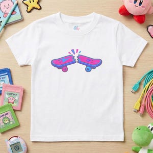 May include: White t-shirt featuring a graphic of a broken skateboard in pink and blue. The skateboard has blue wheels and a lightning bolt design. The shirt is surrounded by video game accessories and toys.