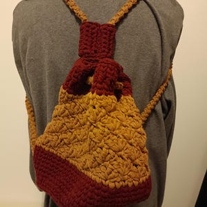 May include: A handmade crochet backpack in shades of burgundy and golden yellow. The bag has a drawstring closure and adjustable straps. The bottom and top section are burgundy, while the main body is golden yellow. The straps are a lighter brown.
