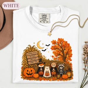 May include: A white t-shirt featuring a graphic print of a fox, a ghost, a bear, and a raccoon dressed in Halloween costumes. The design includes a wooden sign with text, a pumpkin, a crescent moon, bats, and an autumn tree with orange leaves.