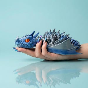 Może przedstawiać: Niebiesko-srebrna figurka smoka z pomarańczowymi oczami. Smok trzymany jest w dłoni na jasnoniebieskim tle. Smok ma szczegółowe łuski i skrzydła i jest przedmiotem o tematyce fantasy.