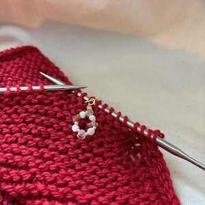 Puede incluir: Primer plano de un proyecto de tejido rojo con agujas de tejer plateadas. Un marcador de puntada dorado con cuentas blancas, marrones y rosas está unido al tejido. El proyecto está en curso, con filas de puntadas de tejido visibles.