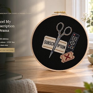 May include: A round wooden embroidery hoop containing black fabric with a cross-stitch design. The design features a pair of scissors, a remote control, and a ticket with an X, alongside the text "SUBSCRIPTION".