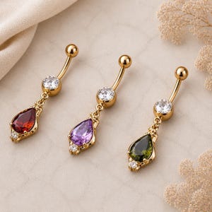May include: Three gold-coloured belly button rings with teardrop-shaped gemstones in red, purple, and green. Each ring features a clear, round gem and small accent stones. The jewellery is displayed on a neutral background.