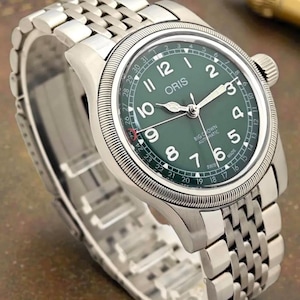 Może przedstawiać: Stalowy zegarek na rękę z zieloną tarczą. Zegarek posiada srebrną bransoletę, wyświetlacz daty oraz nazwę marki "ORIS". Zegarek ma klasyczny design z teksturowaną ramką i koronką.